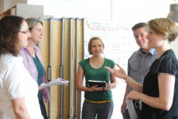 Foto von 5 Personen die eine Unterhaltung führen, drei Personen haben einen Block in der Hand