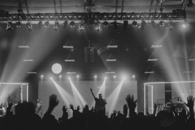 Foto in schwarz weiß von einem Konzert mit Blick auf die Bühne, wo ein Mann steht der seine Hand nach oben hebt