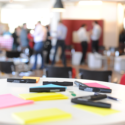 Im Vordergrund ein Tisch mit Stiften, Aufklebern und Post-Its, also Moderationsmaterial, im Hintergrund verschwommen mehrere Personen und Stühle.